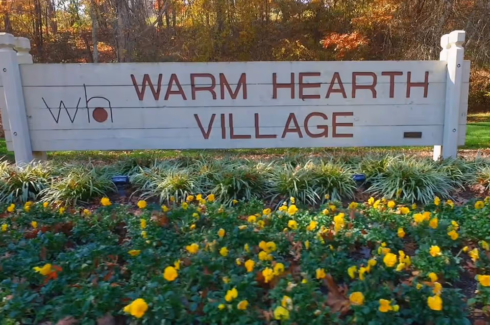 A wooden entrance sign reading 'WARM HEARTH VILLAGE' with yellow flowers and greenery in front and trees behind.