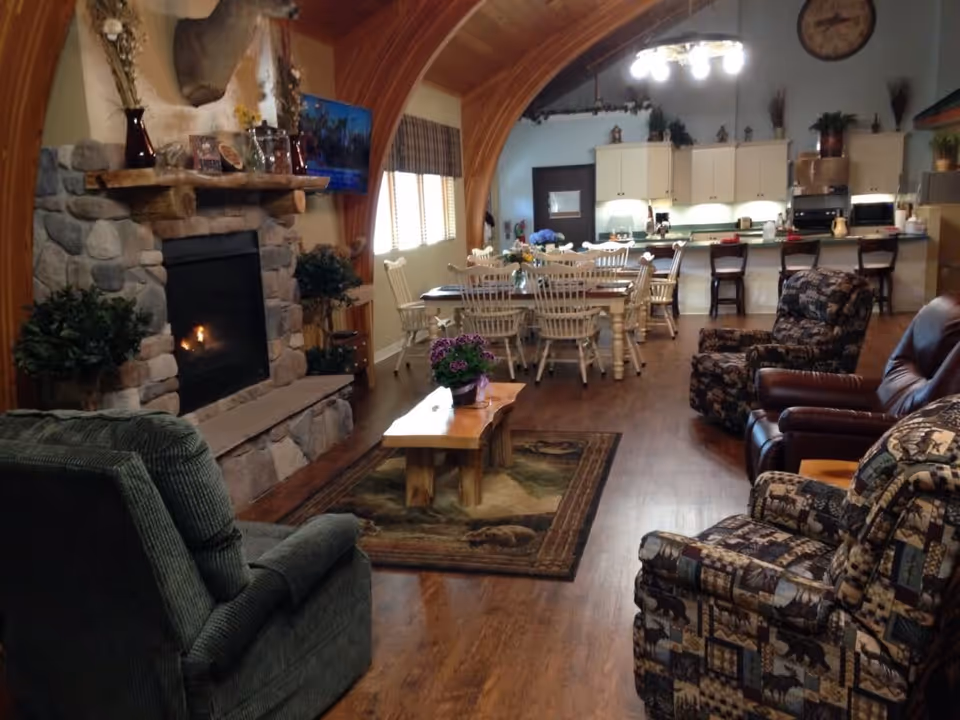 Cozy interior of a living and dining area with wooden floors and a stone fireplace on the left. The room features several upholstered armchairs, a wooden coffee table with a potted plant, a dining table with white chairs, and a kitchen area with white cabinets and bar stools in the background. The ceiling has a wooden arch design and a chandelier light fixture.