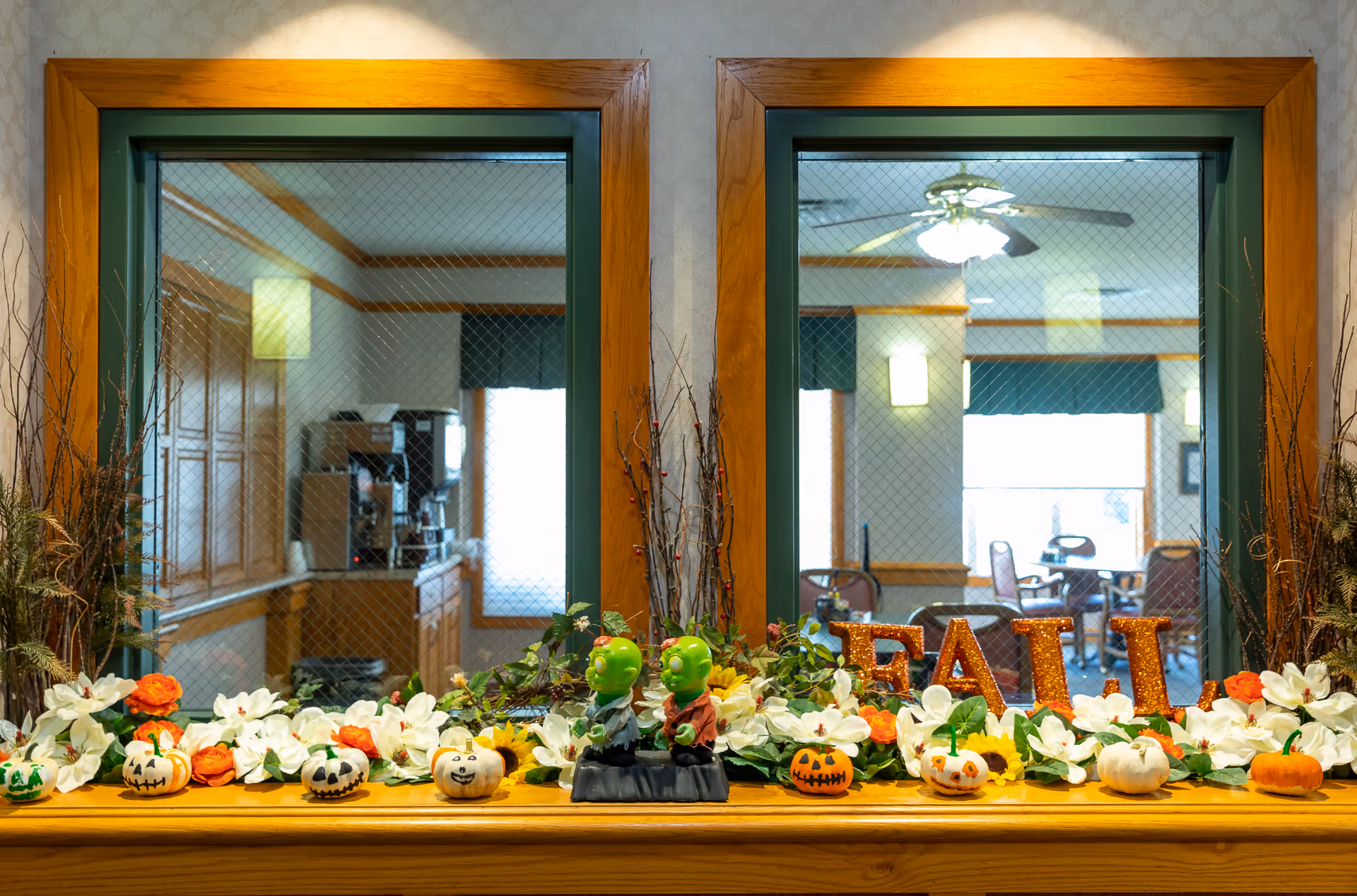 A wooden ledge decorated with artificial white and orange flowers, small decorative pumpkins with faces, two green figurines, and glittery letters spelling 'FALL'. Behind the ledge are two windows with wire mesh, showing an interior room with tables, chairs, and ceiling fans.