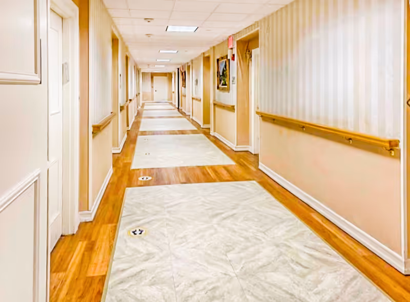 A long, well-lit hallway in a senior living facility with wooden handrails on both sides, light-colored walls with subtle vertical stripes, and a combination of wood and tile flooring. Several doors and framed pictures are visible along the corridor.
