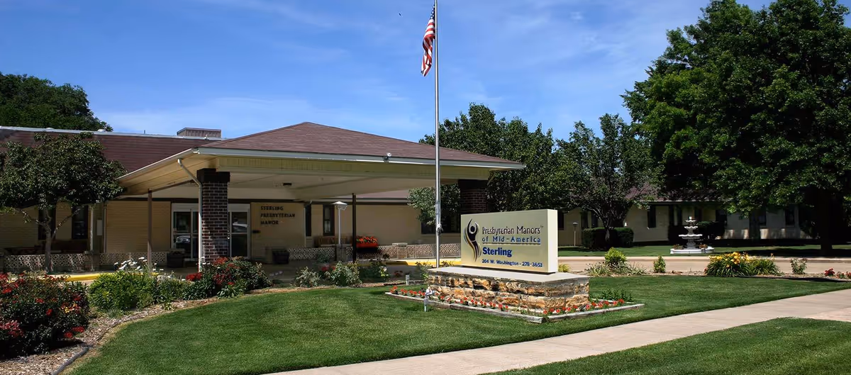 Front entrance of Sterling Presbyterian Manor showing a covered drop-off, flagpole, entrance sign, landscaped lawn, and fountain.