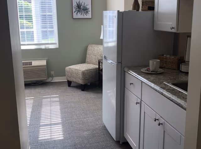 View of a small kitchen area with white cabinets and a granite countertop, a white refrigerator, and a cup on a plate. In the background, there is a window with blinds, a patterned armchair, a small side table, and a wall decoration with botanical artwork.