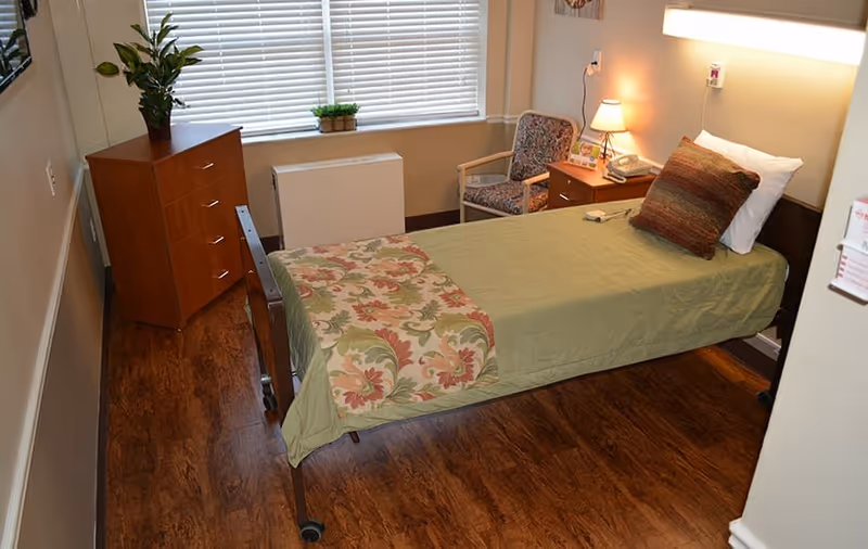 A single bed with green bedding and a floral throw blanket in a nursing facility room. Next to the bed is a wooden nightstand with a lamp, telephone, and a floral-patterned chair. A wooden dresser with a plant on top is against the wall under a window with blinds. The floor is wood, and the room has neutral-colored walls.