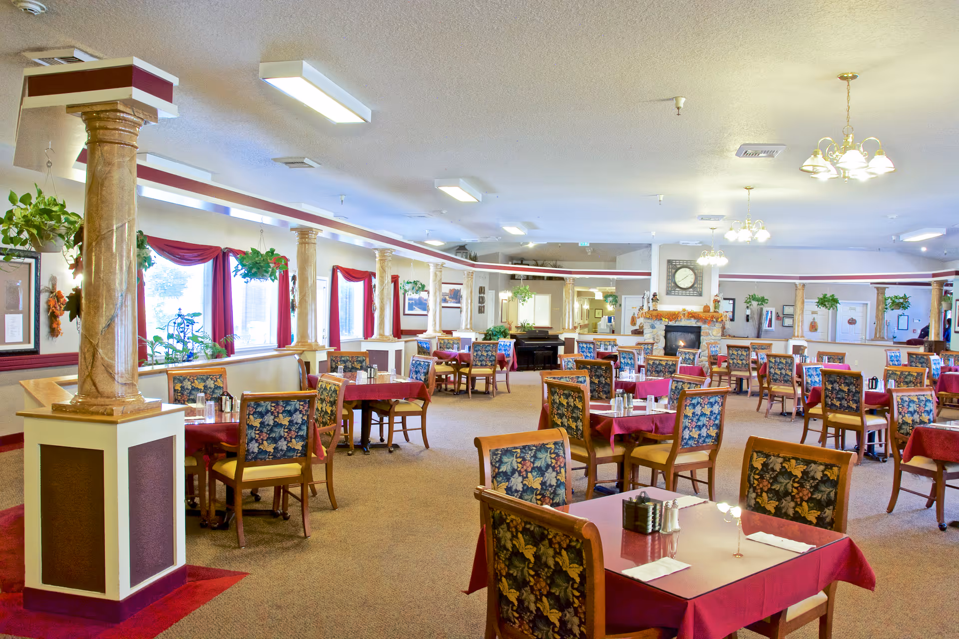A spacious dining room with multiple tables covered in red tablecloths and surrounded by wooden chairs with floral upholstery. The room features large windows with red curtains, decorative columns, hanging plants, and chandeliers. A fireplace with a clock above it is visible in the background.
