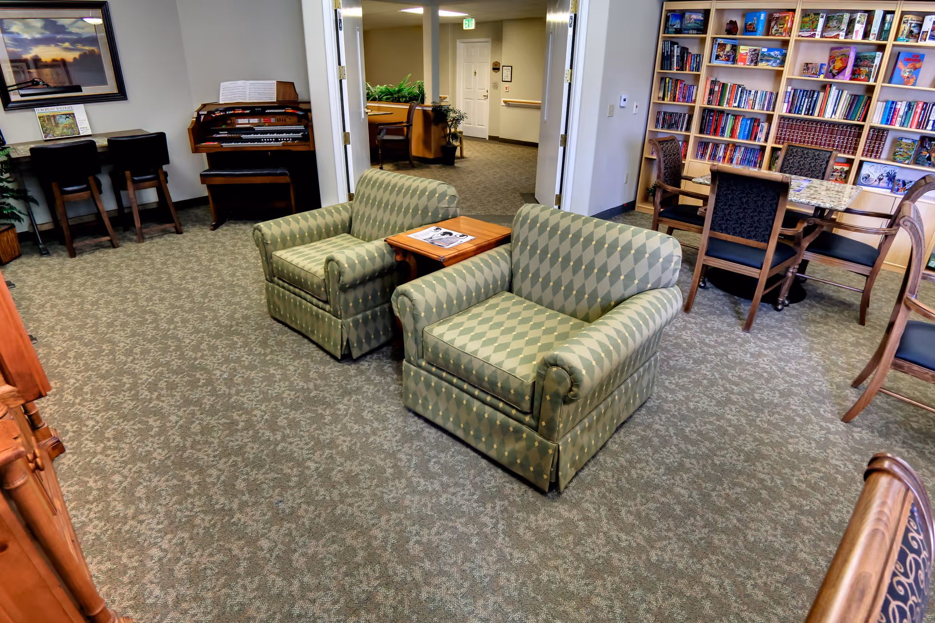 Two patterned armchairs and a side table in a carpeted common room with bookshelves, tables, and a small organ.
