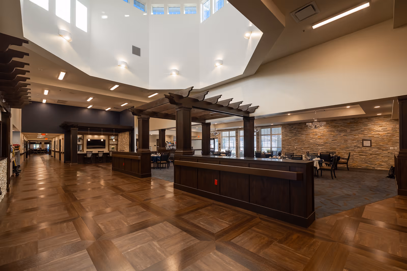 Spacious interior of a senior living facility featuring a large open area with wooden flooring and dark wood architectural accents. The space includes a dining area with tables and chairs set for meals, stone accent walls, and large windows allowing natural light. The ceiling is high with multiple wall-mounted lights and a skylight above.