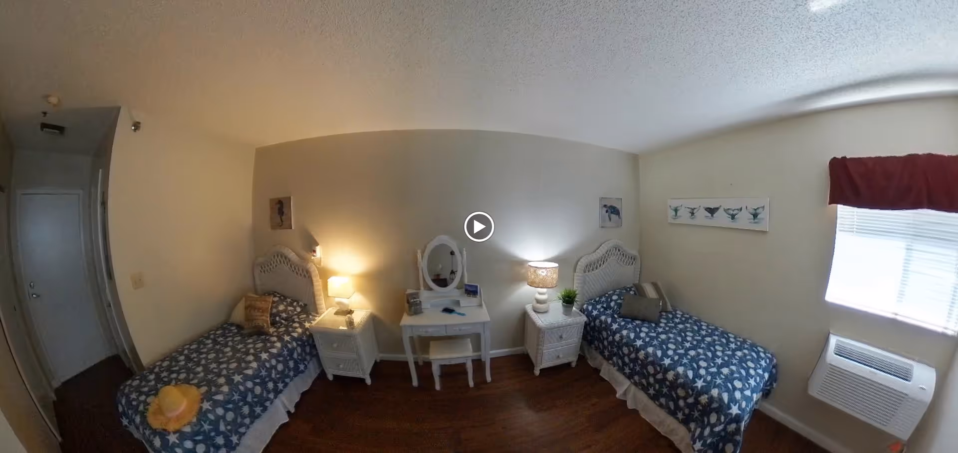A bedroom with two twin beds, each with a blue and white starfish patterned bedspread. Between the beds is a small white vanity table with a mirror and a white chair. Each bed has a white nightstand with a lamp. There is a window with a red valance on the right wall and an air conditioning unit below it. The floor is wooden, and the walls are painted light beige. The room has a door on the left side.