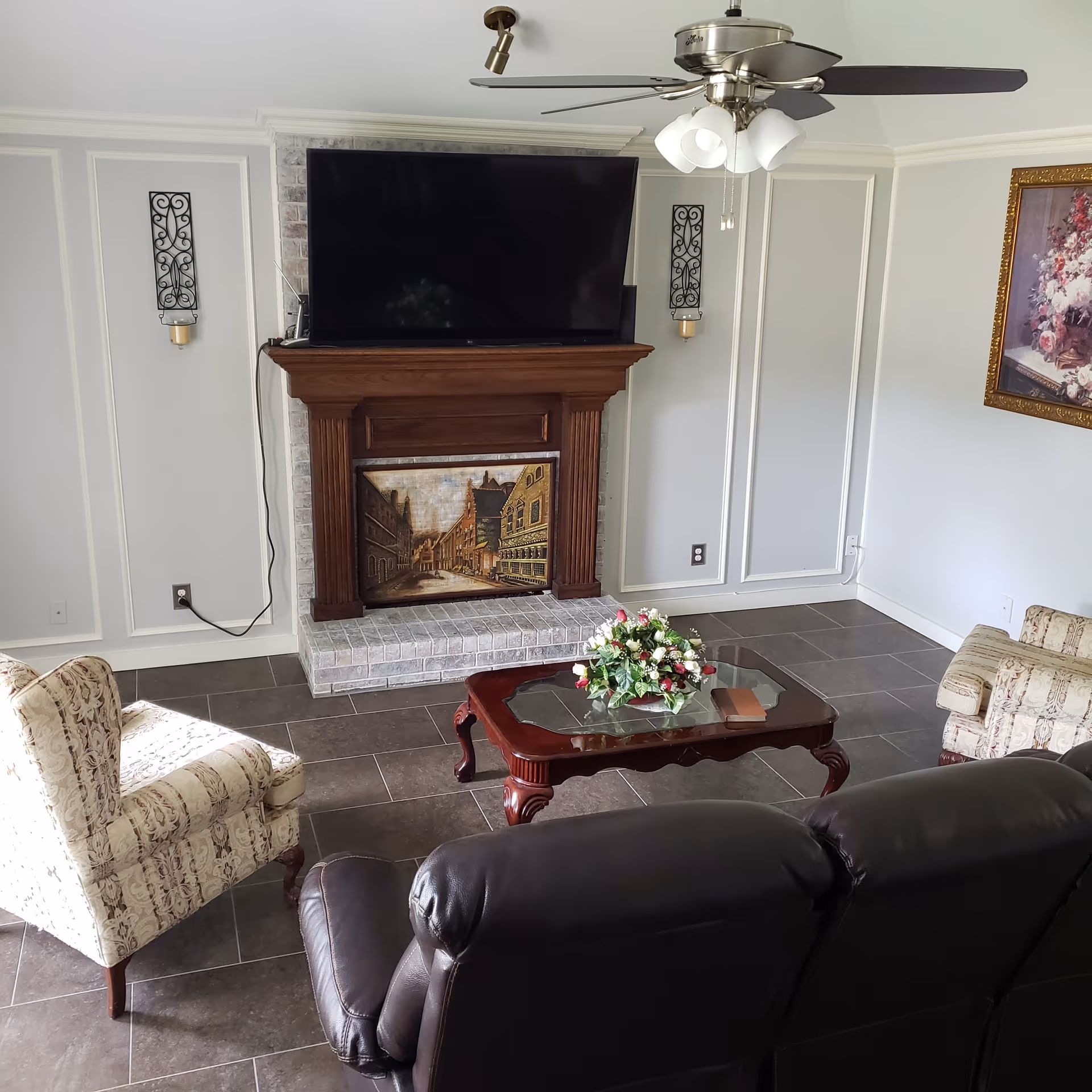 Cozy living room with a large TV mounted above a wooden fireplace mantel, a coffee table with a floral centerpiece, and upholstered seating.