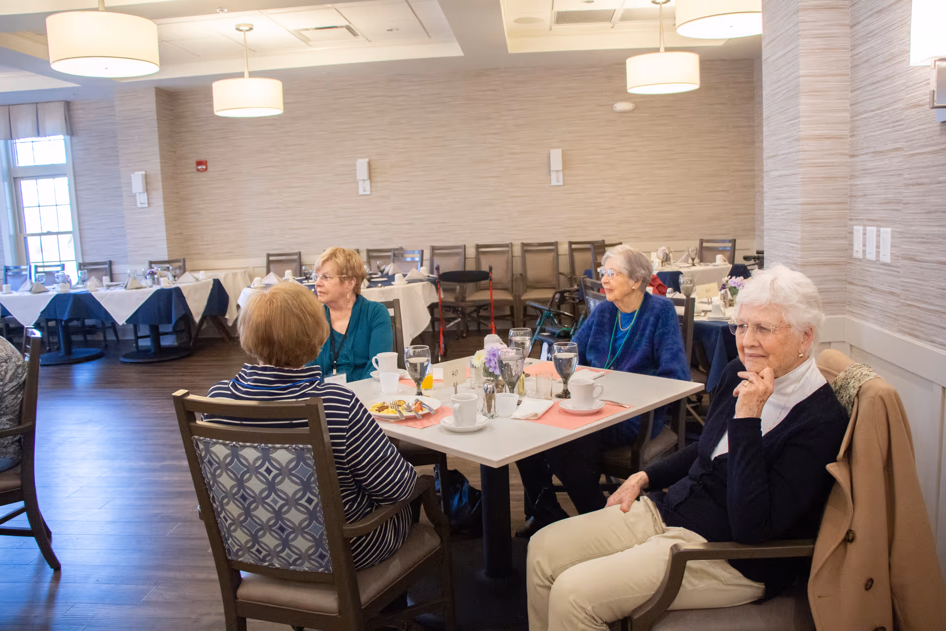 Four elderly women sitting around a table in a dining room, engaged in conversation. The table is set with cups, glasses, and plates. The room has wooden flooring, multiple tables with white and blue tablecloths, and large windows letting in natural light.