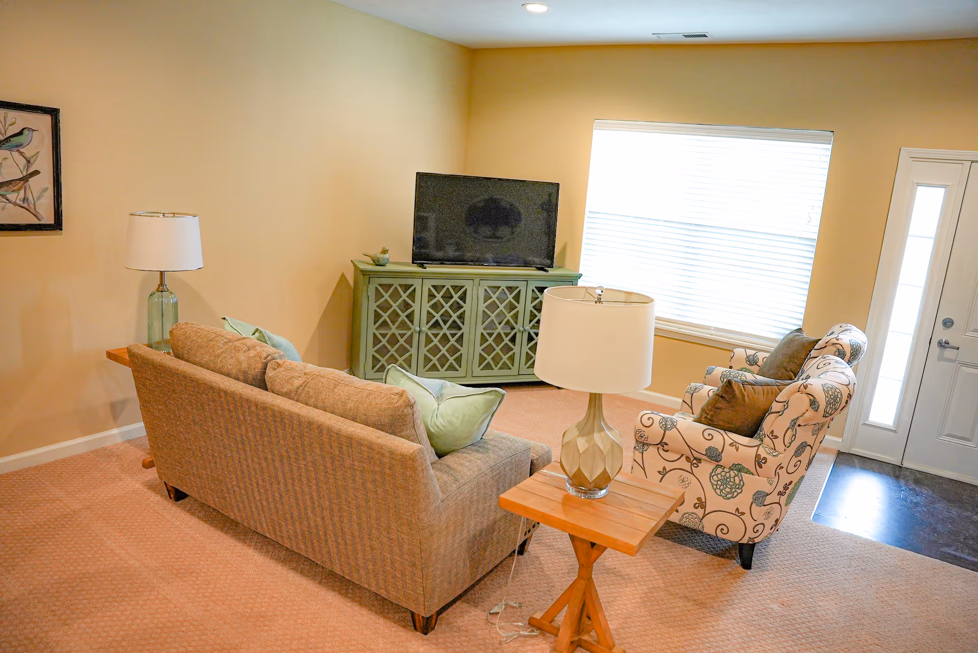 A cozy living room with a beige couch adorned with light green pillows, a patterned armchair with brown cushions, a wooden side table with a lamp, a green TV stand with a flat-screen television, a window with blinds, and a door with glass panels. The walls are painted a soft beige color and the floor is carpeted.