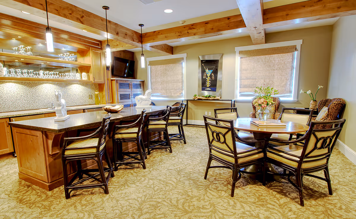 A cozy dining and lounge area with a large kitchen island featuring several high chairs, wooden cabinetry, and a granite countertop. The room has warm lighting with pendant lights hanging from a wooden beam ceiling. There is a round wooden table with four cushioned chairs, a vase with flowers, and two wicker armchairs near the windows with woven blinds. A wall-mounted TV and decorative artwork are also visible.