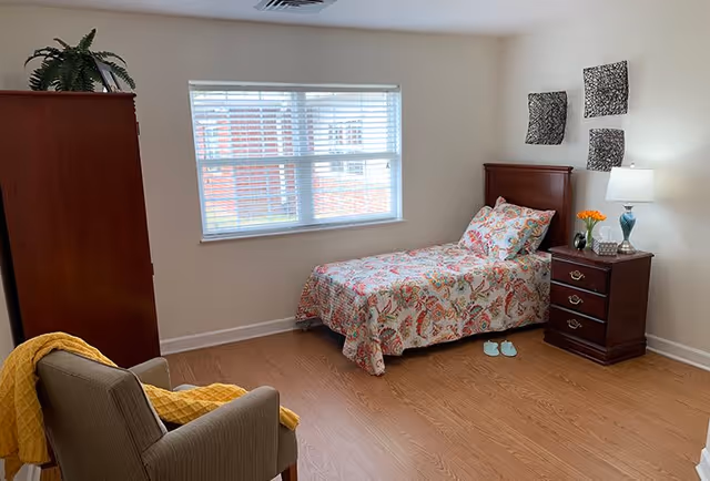 A simple bedroom with a single bed covered in a colorful floral bedspread, a wooden nightstand with a lamp and a small vase of flowers, a wooden wardrobe, a comfortable armchair with a yellow blanket draped over it, and a window with white blinds letting in natural light. The walls are light-colored with three decorative square wall hangings above the bed.