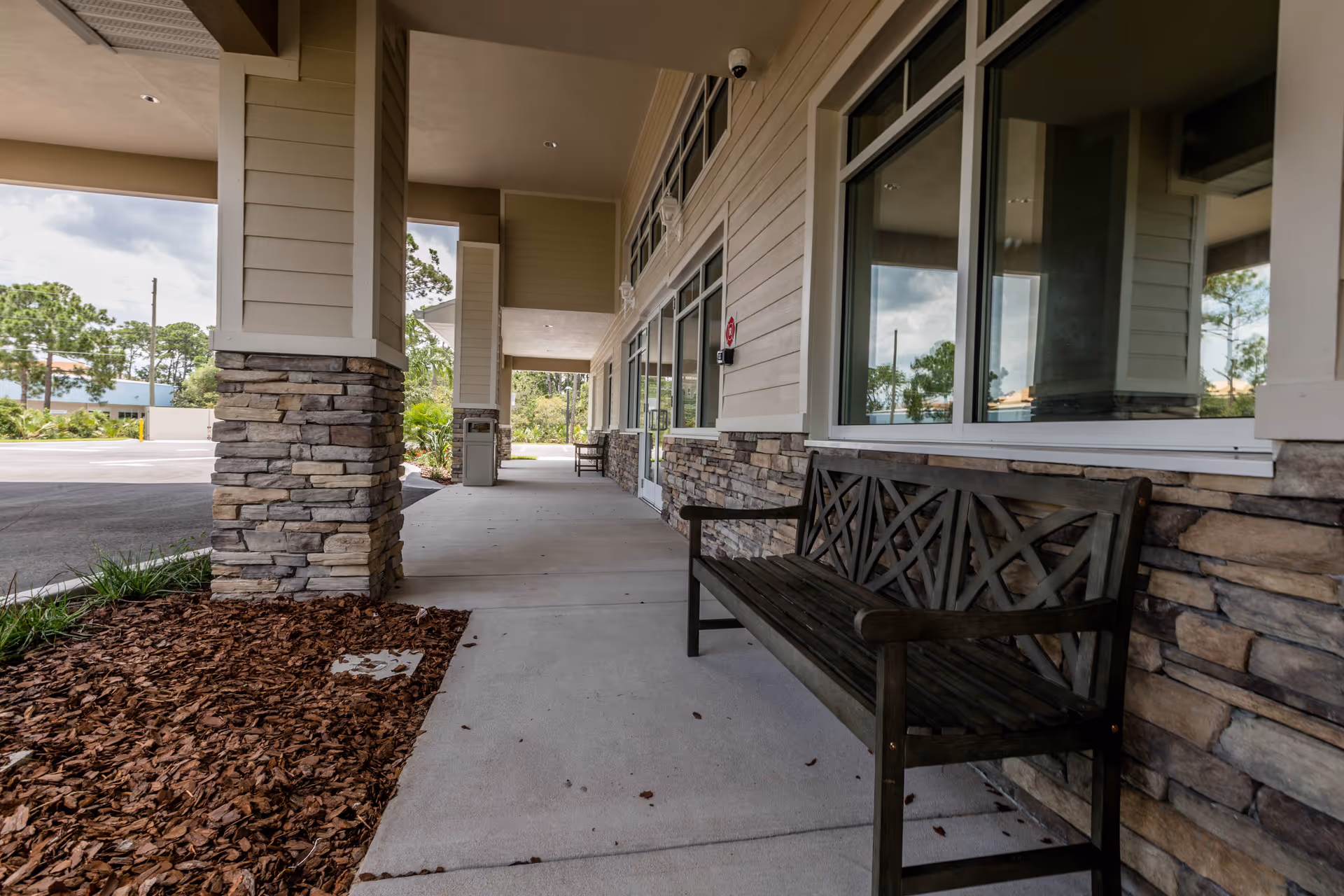 Covered outdoor walkway with stone pillars and a wooden bench along the wall of a building, with windows reflecting the sky and trees in the background.