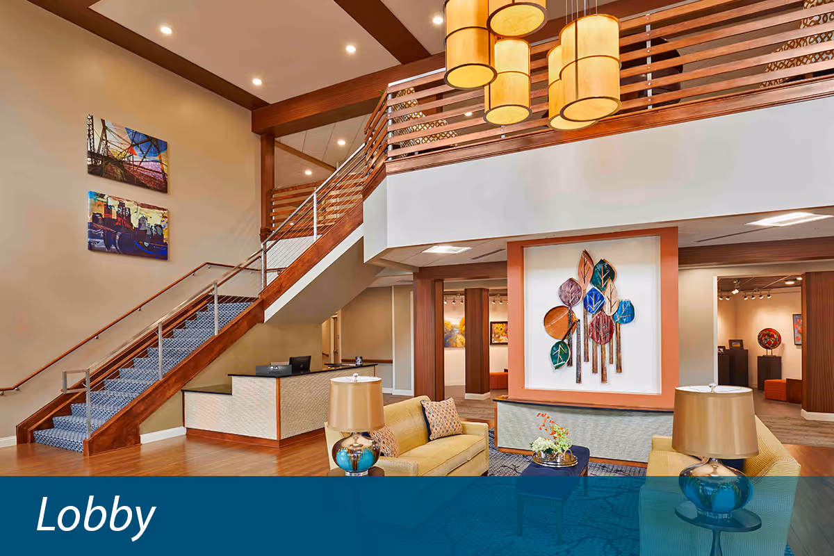 A spacious and well-lit lobby area with a modern staircase featuring wooden railings and blue carpeted steps. The lobby has comfortable seating with yellow sofas, a coffee table with a flower arrangement, and two lamps with beige shades. The walls are decorated with colorful artwork, including a large piece with abstract leaf designs. The ceiling has recessed lighting and hanging cylindrical light fixtures.
