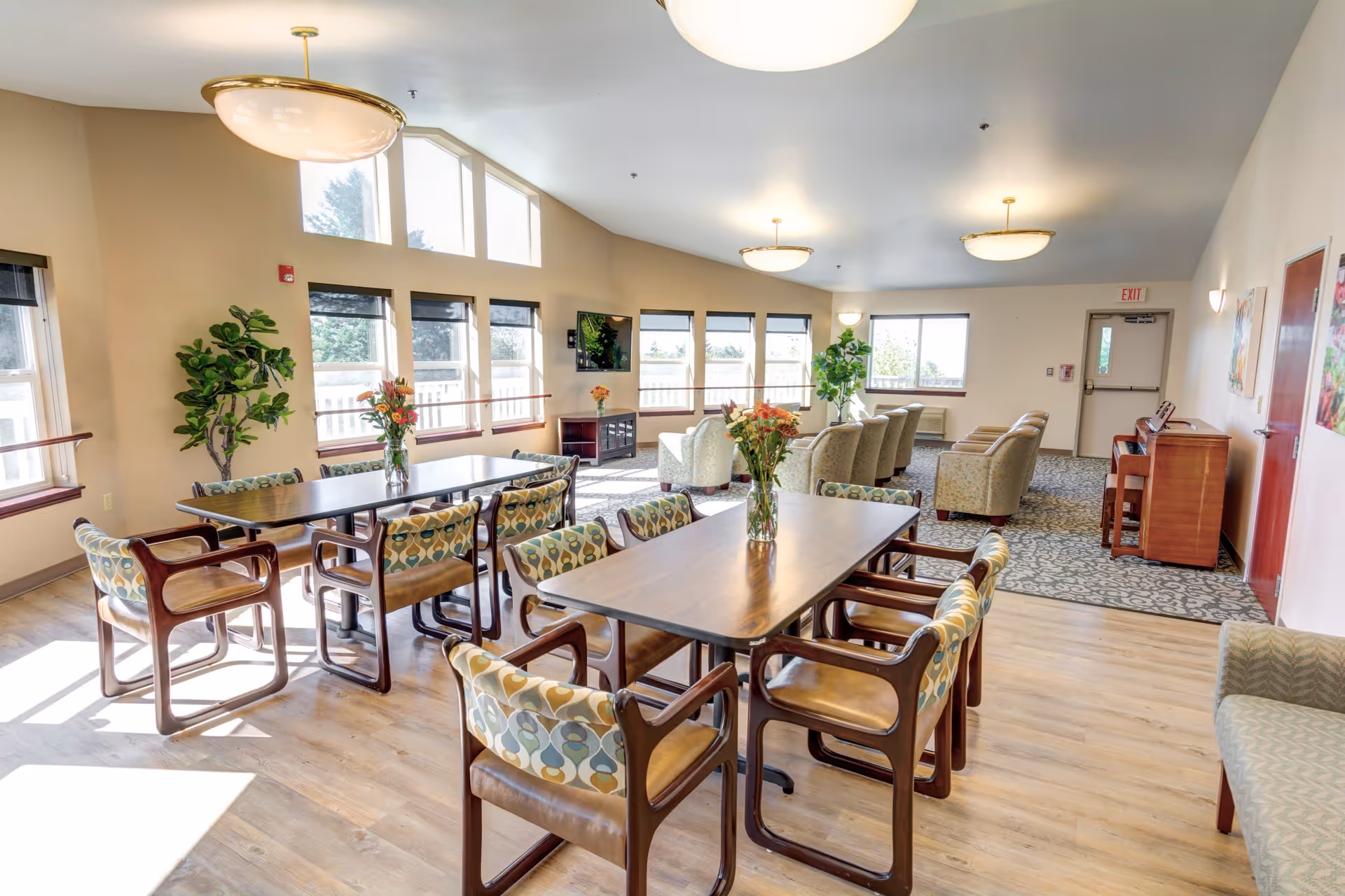 A bright and spacious common area in a senior living facility with large windows letting in natural light. The room features two wooden tables with patterned cushioned chairs around them, vases with flowers on the tables, several armchairs arranged in a seating area near the windows, a piano against the wall, and potted plants. The flooring is a combination of wood and carpet, and the ceiling has round light fixtures.