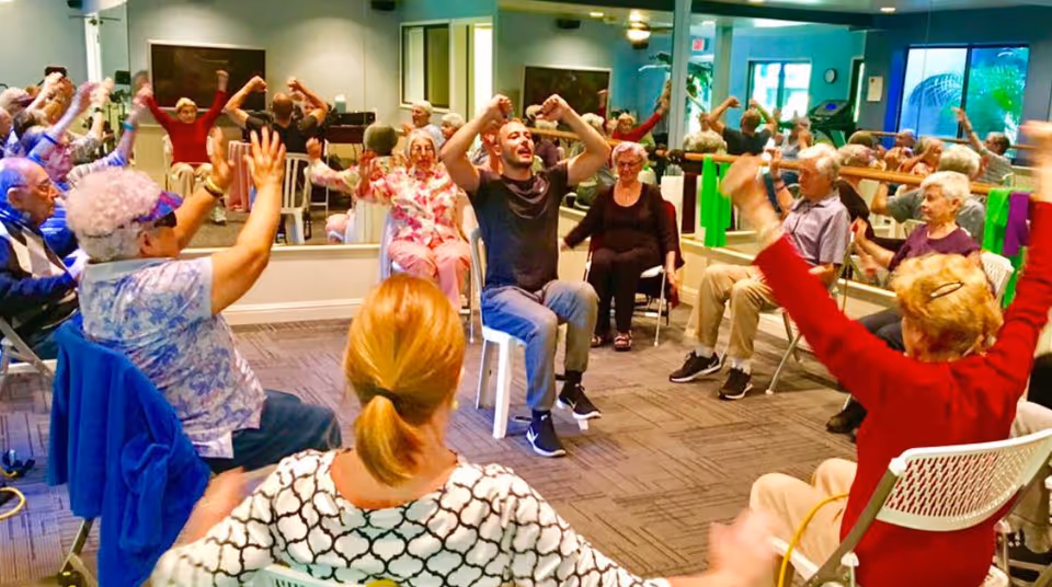 A group of elderly people sitting in chairs arranged in a circle in a room, participating in a seated exercise or stretching activity led by a younger instructor in the center. The room has mirrors on the walls and exercise bands hanging on a barre.