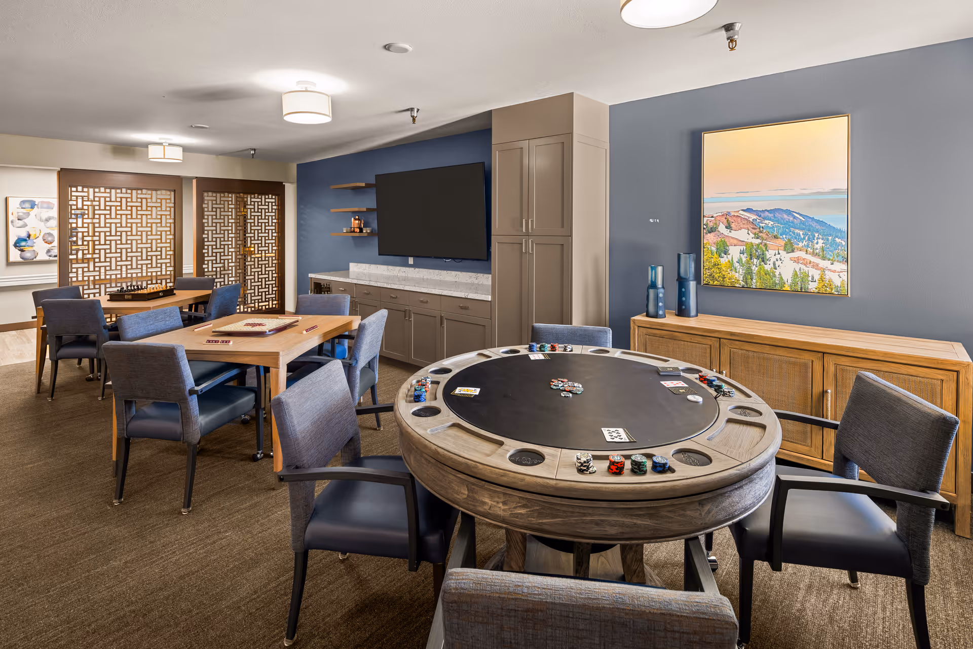 A senior living community game room with a round poker table in the foreground, surrounded by six chairs. The table has poker chips and cards on it. In the background, there are two rectangular tables with chairs, one with a chessboard on it. The room features a blue accent wall with a mounted flat-screen TV, cabinetry, and a landscape painting. The floor is carpeted, and the room is well-lit with ceiling lights.