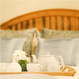 A wooden tray on a bed with a white teapot, cups, a small pitcher, a sugar bowl, and a small vase with white flowers. The bed has a wooden headboard and pillows with blue and patterned pillowcases.