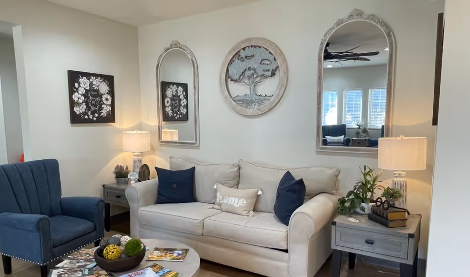 A cozy living room with a beige sofa adorned with two dark blue pillows and a beige pillow with the word 'home'. There is a blue armchair to the left and two gray side tables with lamps and decorative plants on either side of the sofa. Above the sofa are two tall mirrors and a round wall decoration featuring a tree. A coffee table in front of the sofa holds a bowl with decorative balls and some magazines.