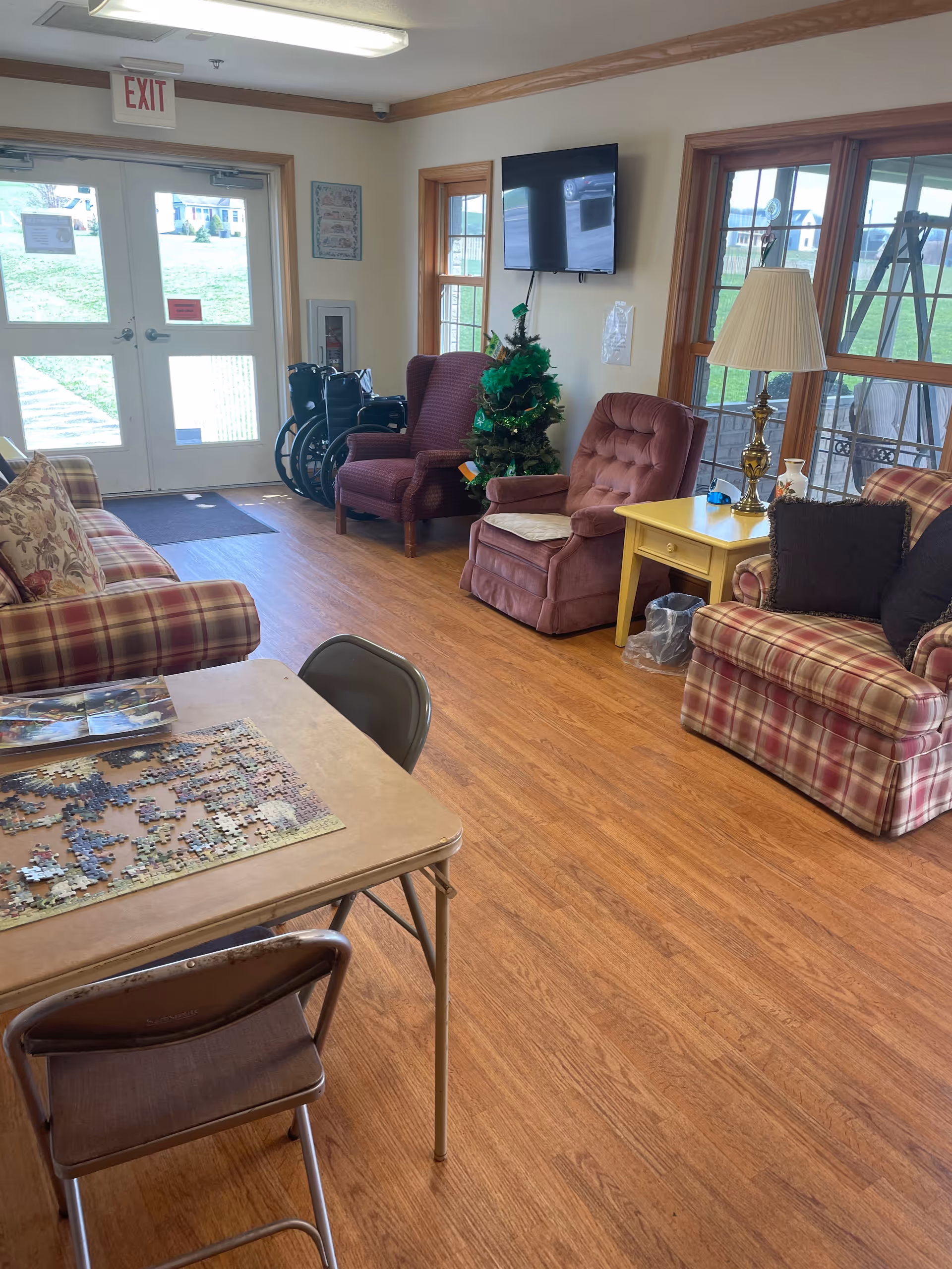 A cozy assisted living common area with wooden flooring, two plaid sofas with cushions, two armchairs, a small decorated Christmas tree, a yellow side table with a lamp, a wall-mounted TV, and a table with a partially completed jigsaw puzzle and folding chairs. Large windows and a glass door provide natural light and a view of the outside grassy area.