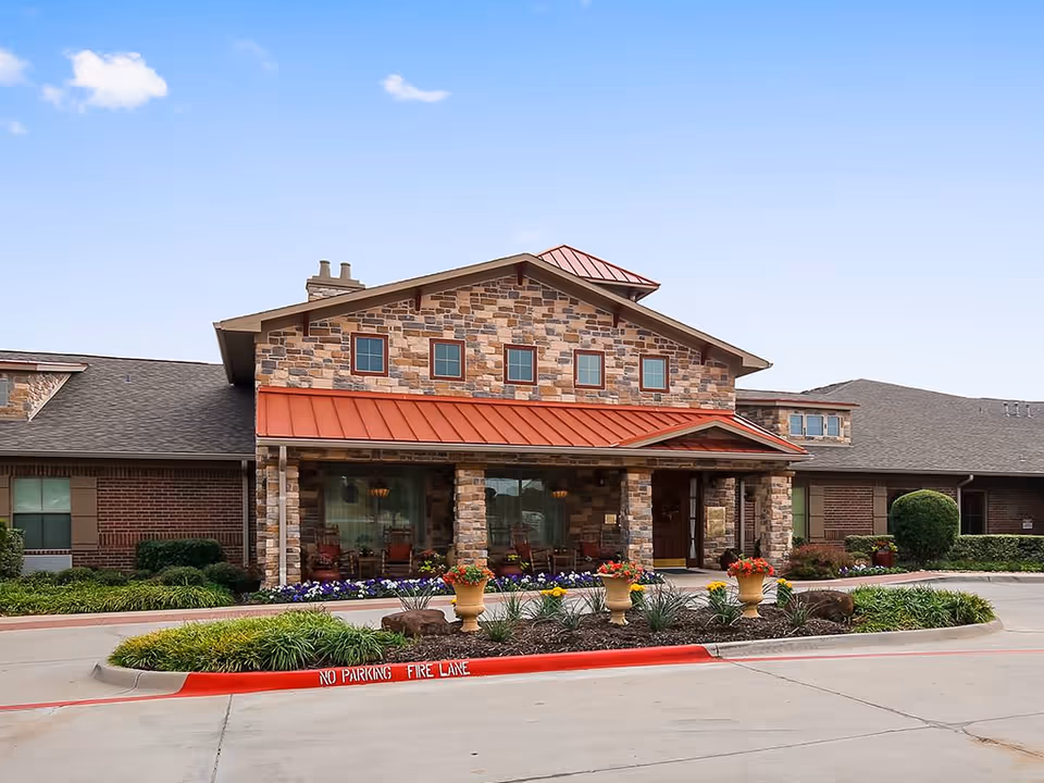 Front exterior view of a senior living facility building with a stone and brick facade, a red metal roof over the entrance, and a landscaped area with flowers and plants in front. The sky is clear and blue.