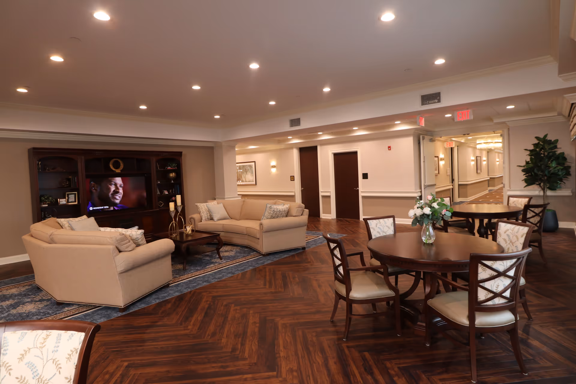 A spacious senior living facility common area with two beige sofas arranged around a wooden coffee table on a blue patterned rug. A large wooden entertainment center with a TV is against the wall. In the foreground, there are round wooden tables with floral upholstered chairs and a vase with flowers on one table. The room has wood-patterned flooring, recessed ceiling lights, and a hallway with multiple doors and exit signs.