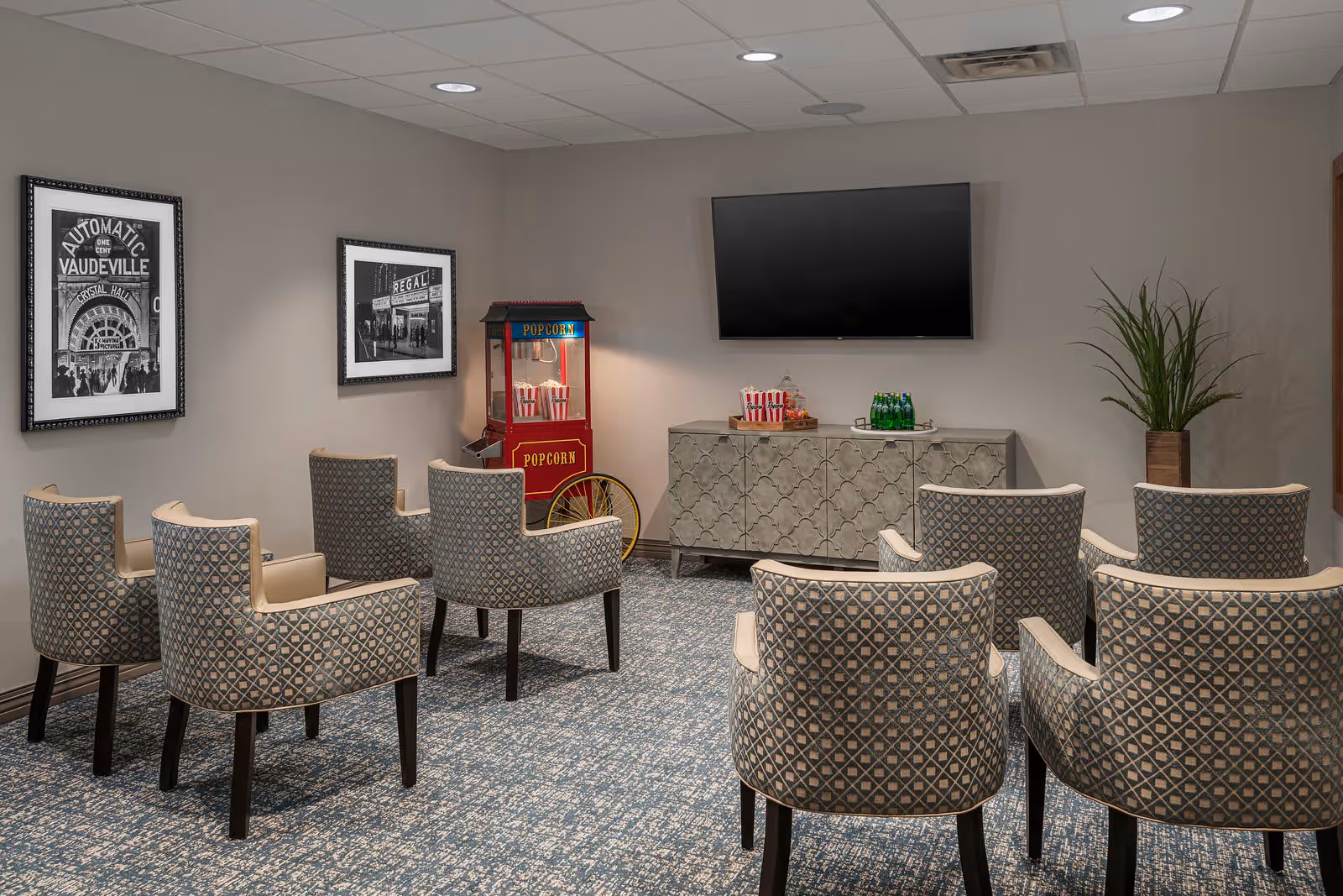 A small theater-style room with eight patterned armchairs arranged in two rows facing a wall-mounted flat screen TV. A vintage-style popcorn machine with popcorn bags is positioned in the corner next to a decorative cabinet holding more popcorn bags and bottled drinks. Two black and white framed photos hang on the wall, and a potted plant is placed in the corner.
