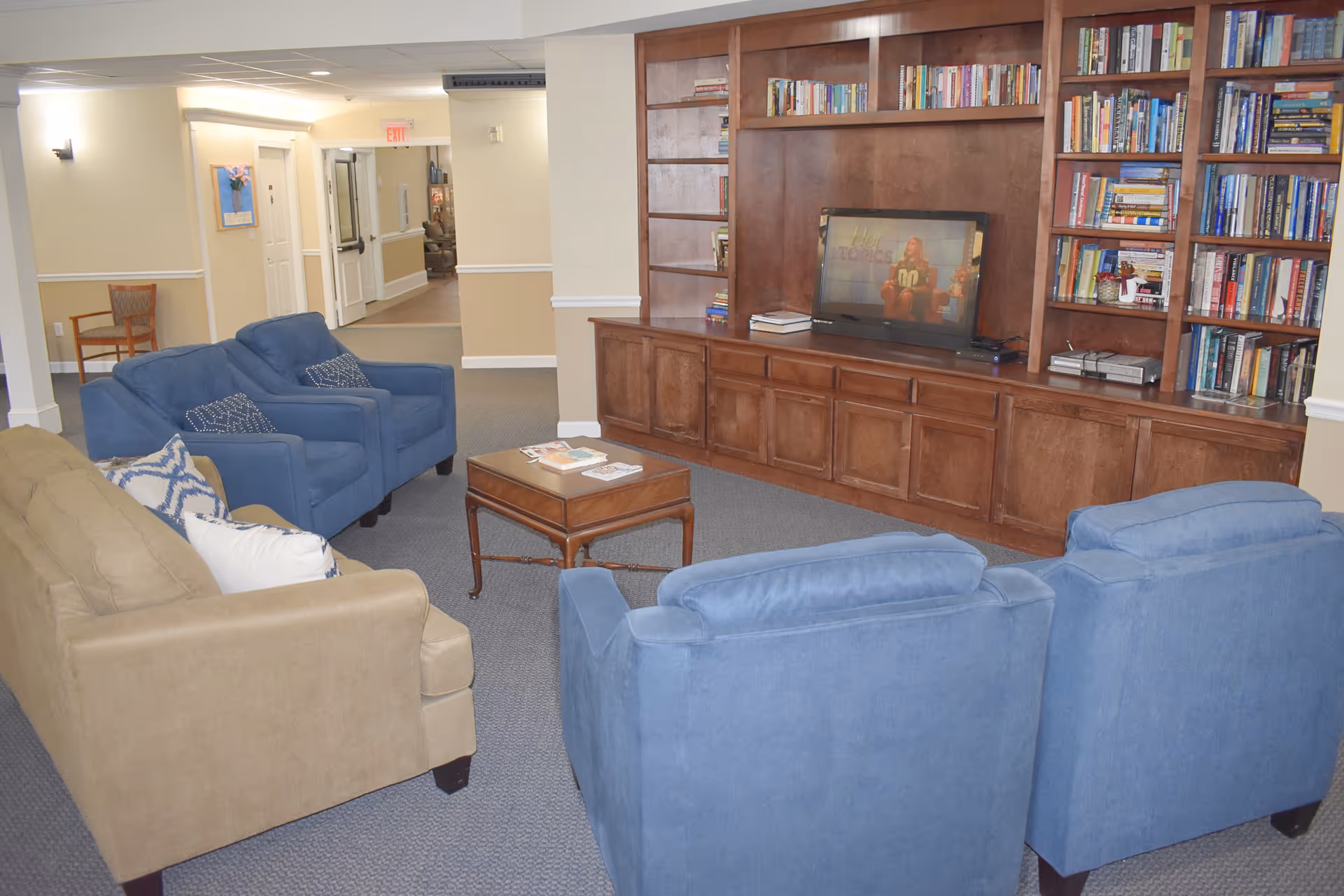 A cozy common area with a beige sofa and four blue armchairs arranged around a wooden coffee table. A large wooden built-in bookshelf and entertainment center with a flat-screen TV is against the wall, filled with books and decorative items. The room has beige walls and carpeted flooring, with a hallway visible in the background.