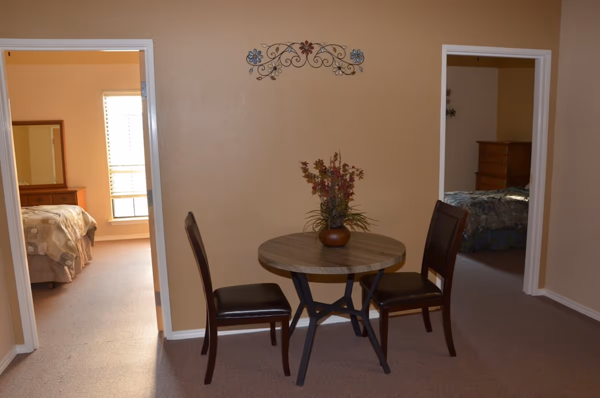 A small round wooden table with a flower vase centerpiece and two dark wooden chairs with black cushions are placed against a beige wall. On the wall above the table is a decorative metal wall art with floral designs. Two doorways on either side of the table lead to bedrooms, each containing a bed and wooden furniture.