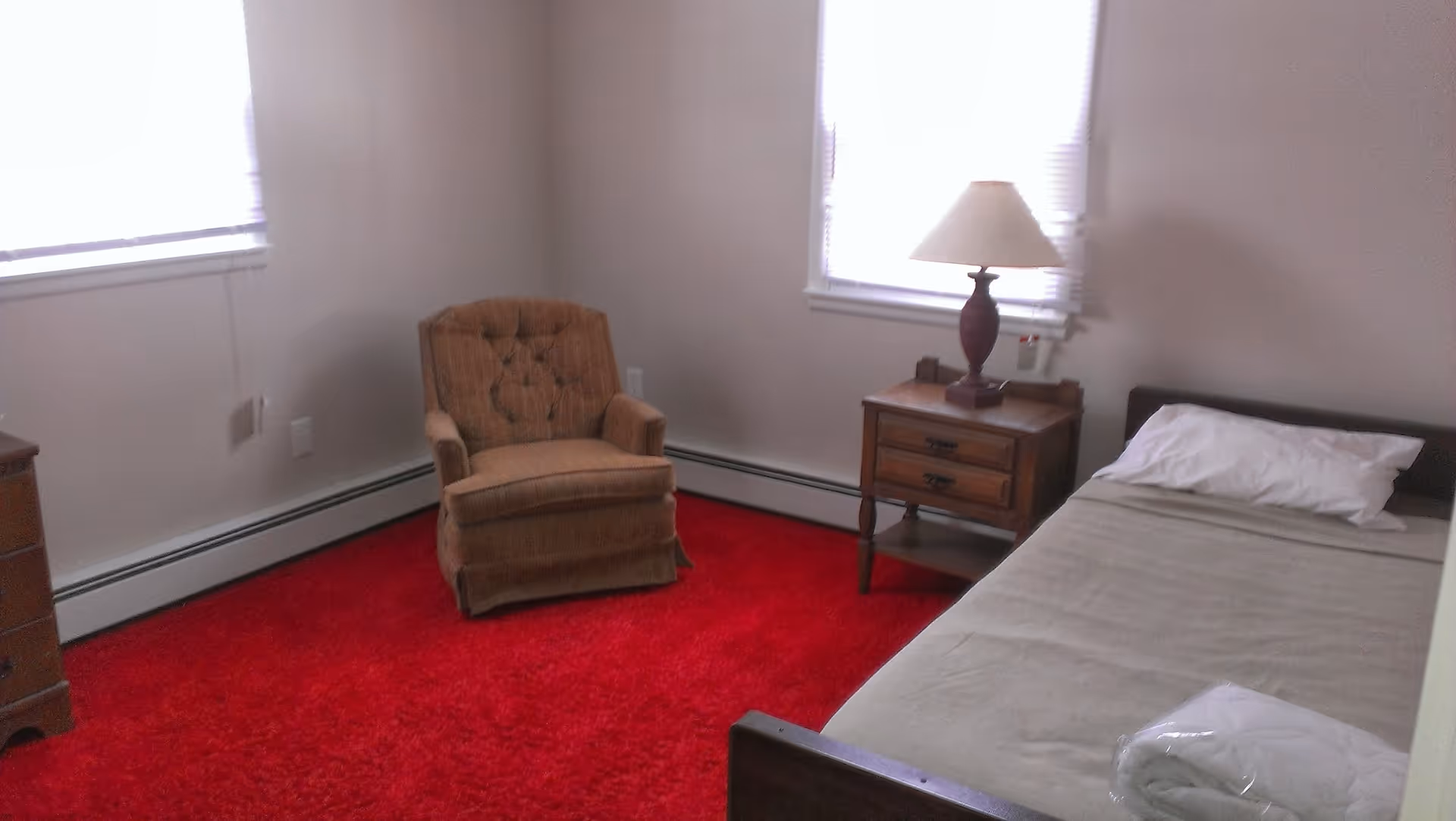 Small bedroom with a single bed, wooden nightstand and lamp, an upholstered armchair, and a bright red carpet beneath two windows.