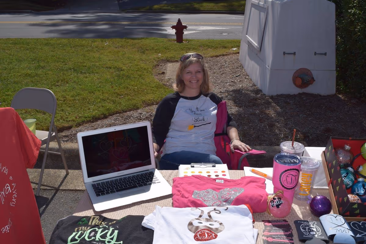 A woman sitting outdoors behind a table displaying various colorful t-shirts, a laptop, drink containers, and decorative items. She is smiling and wearing a white and black raglan shirt with text on it. The setting appears to be a sunny day with grass and a road in the background.