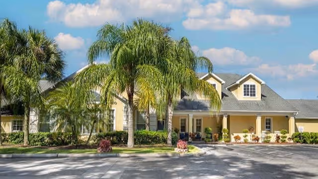 Exterior view of a single-story senior living facility building with a gray roof, beige walls, and a covered entrance. Several palm trees and shrubs are planted in front of the building, and a paved driveway leads to the entrance under a partly cloudy sky.