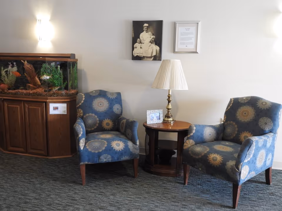 A cozy sitting area with two blue patterned armchairs facing each other, separated by a small wooden side table with a brass lamp and a framed photo. To the left, there is a large fish tank with plants and fish inside. On the wall above the chairs, there is a black and white photo of a nurse holding two children and a framed document.