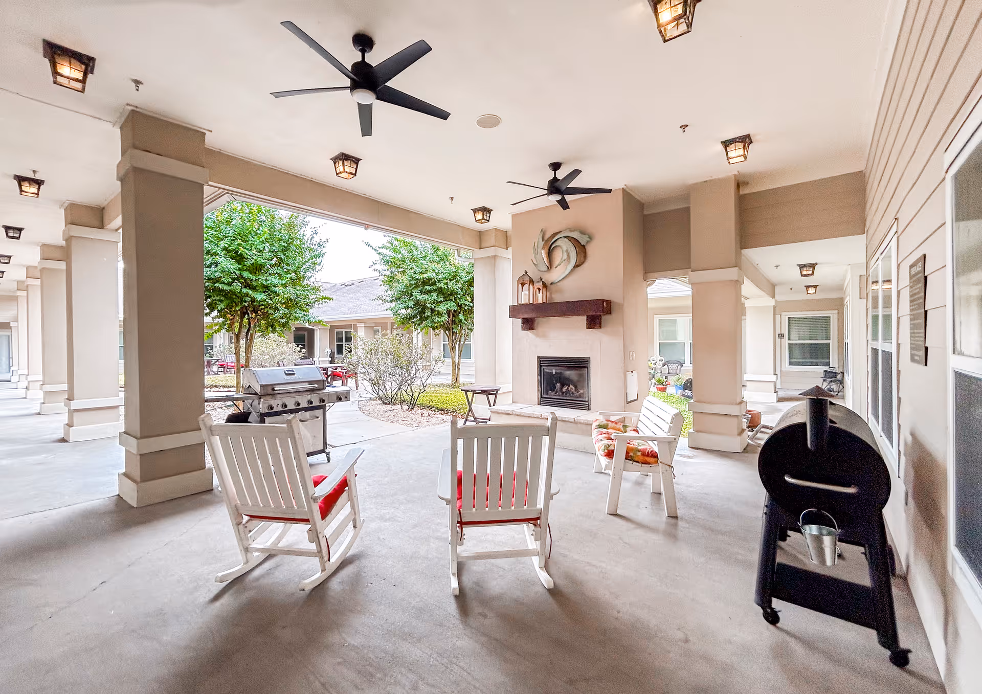 Covered outdoor patio area with white rocking chairs arranged around a fireplace. There are ceiling fans and lantern-style lights on the ceiling. A grill and a smoker are also visible, with trees and a garden area in the background.