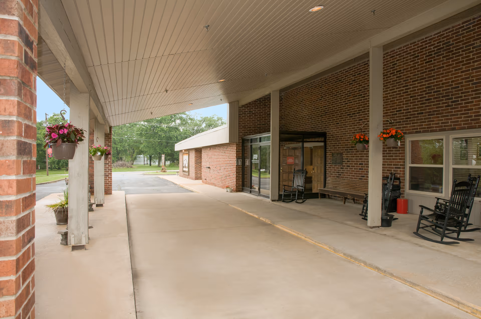 Covered entrance area of a brick building with hanging flower pots, black rocking chairs, and a bench near the glass double doors. Trees and a driveway are visible in the background.