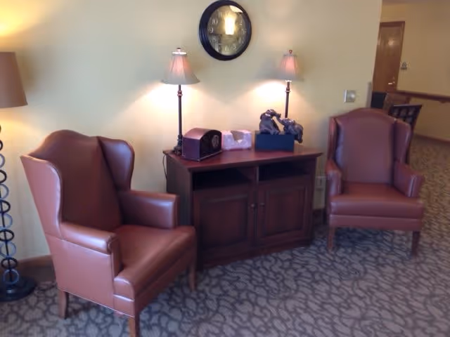 Two leather armchairs flank a wooden console with lamps and a wall clock in a carpeted seating area.