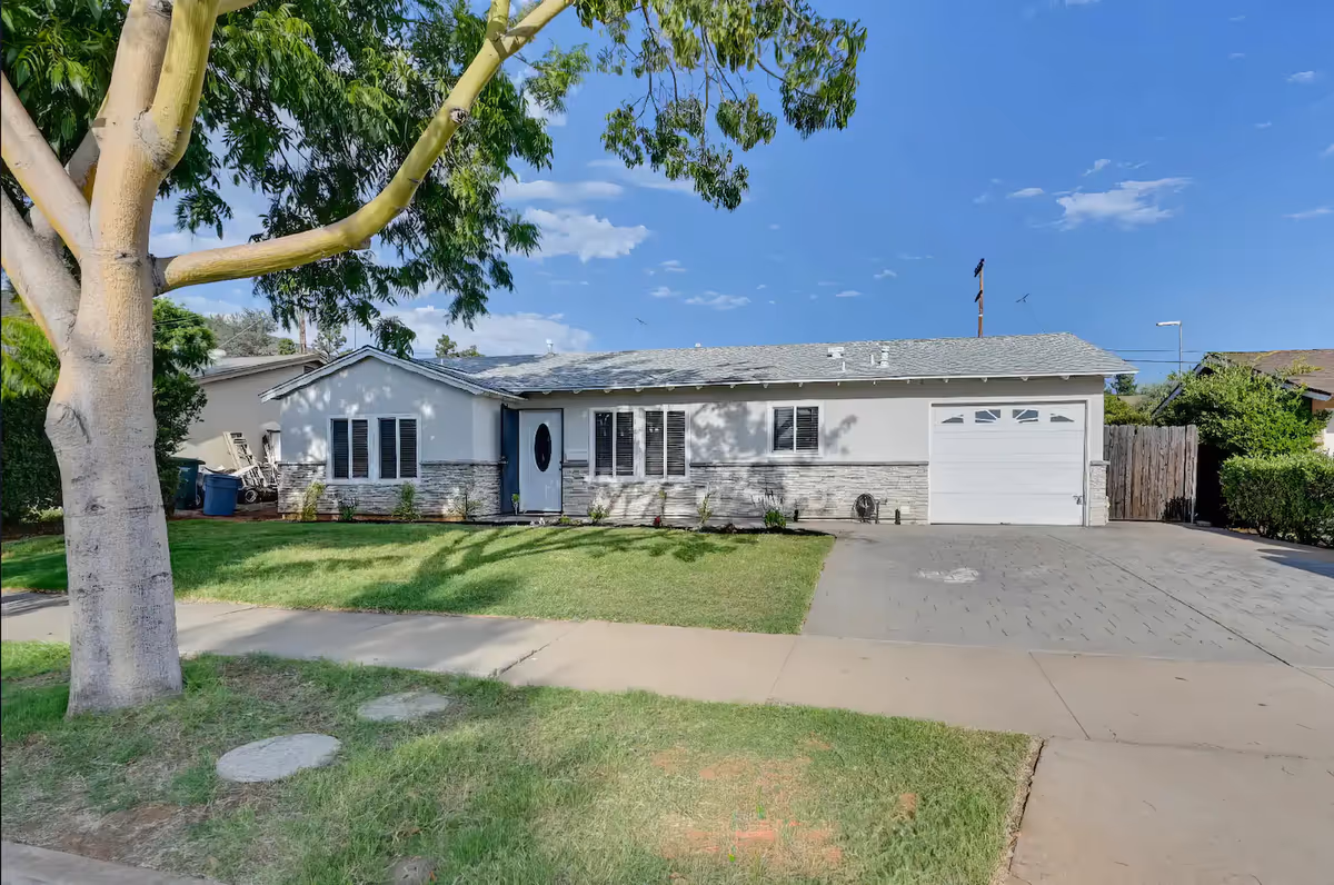 Single-story residential building with a light gray exterior, white garage door, and a front lawn with a large tree. The sky is clear and blue.