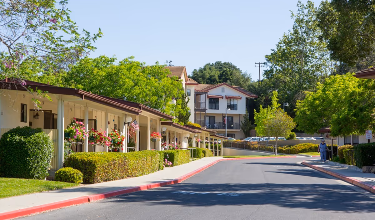 View of a senior living community street with single-story buildings on the left adorned with hanging flower pots and American flags, well-maintained bushes, trees, and a person walking on the sidewalk on the right side under clear blue sky.