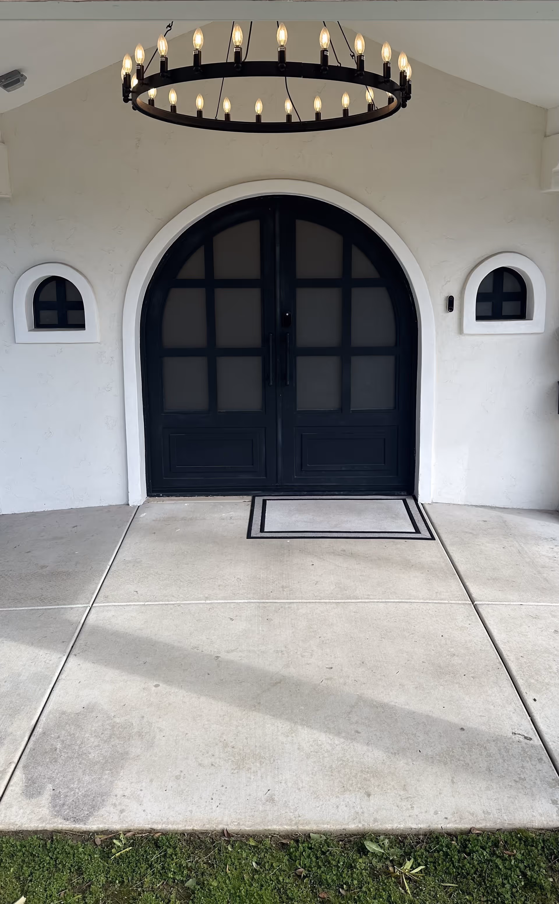 Black double arched doors with frosted glass panels set in a white stucco wall, flanked by two small arched windows. A large circular chandelier with multiple candle-like lights hangs above the entrance. Concrete walkway and green grass are visible in front of the doors.