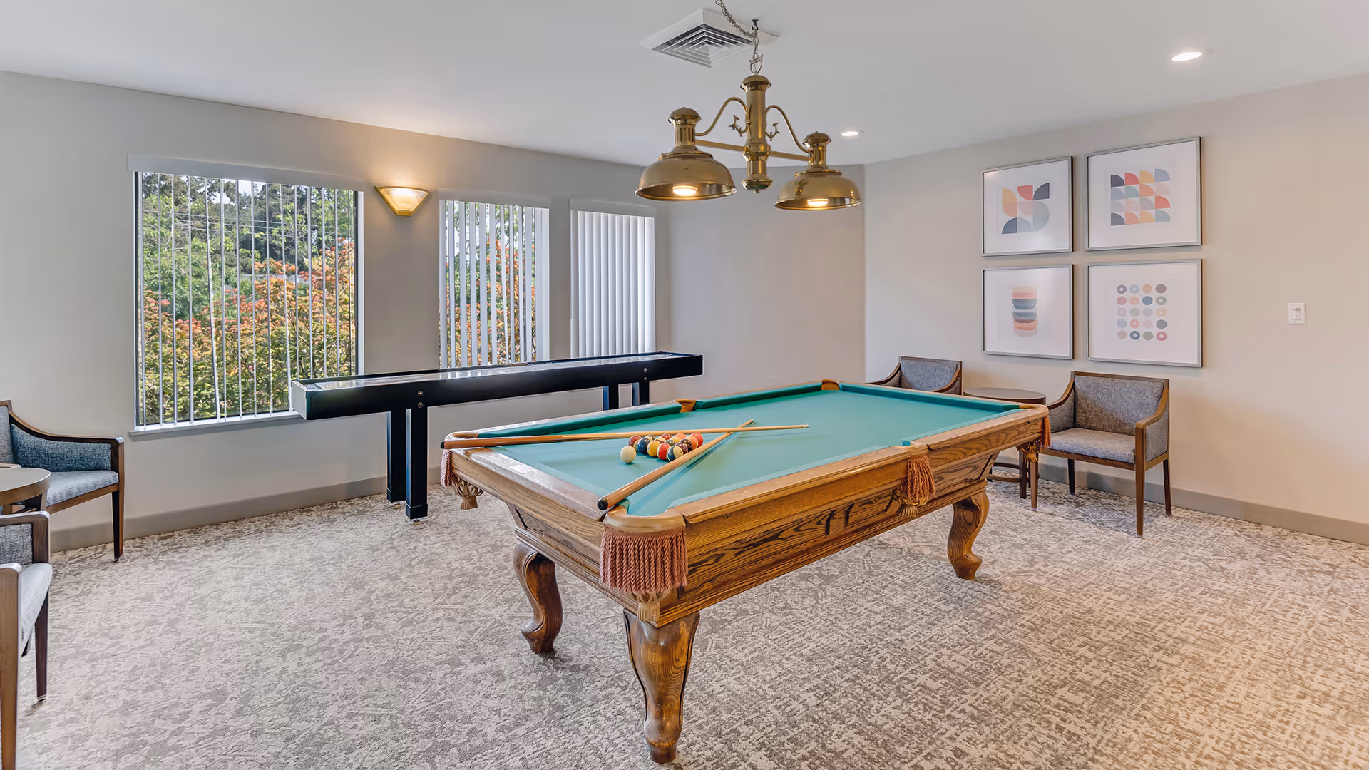 A recreational room with a pool table in the center, equipped with pool balls and cues. The room has carpeted flooring, three framed abstract art pieces on the wall, and several chairs arranged around the room. Large windows with vertical blinds allow natural light to enter, and a shuffleboard table is positioned against the wall beneath the windows. A brass light fixture hangs above the pool table.