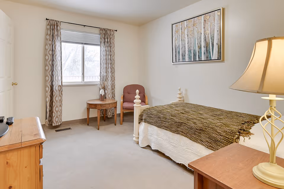 A simple, cozy bedroom with a single bed covered with a brown textured blanket. There is a wooden nightstand with a lamp on the right side of the bed. A wooden dresser with a TV remote is on the left side of the room. A window with patterned curtains lets in natural light. A round wooden table with a vase and a maroon upholstered chair are positioned near the window. A framed painting of birch trees hangs on the wall above the bed.