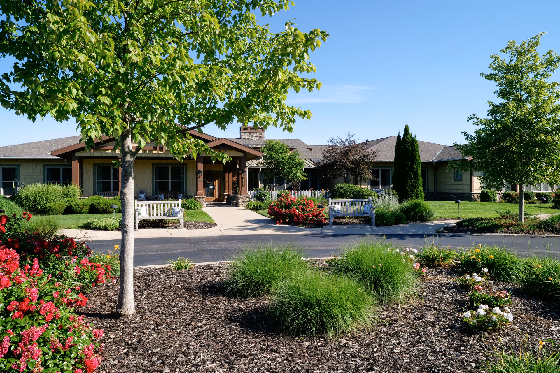 Exterior view of a single-story senior living facility building with a landscaped garden in front, featuring green trees, bushes, colorful flowers, and two white benches along a paved walkway leading to the entrance under a clear blue sky.