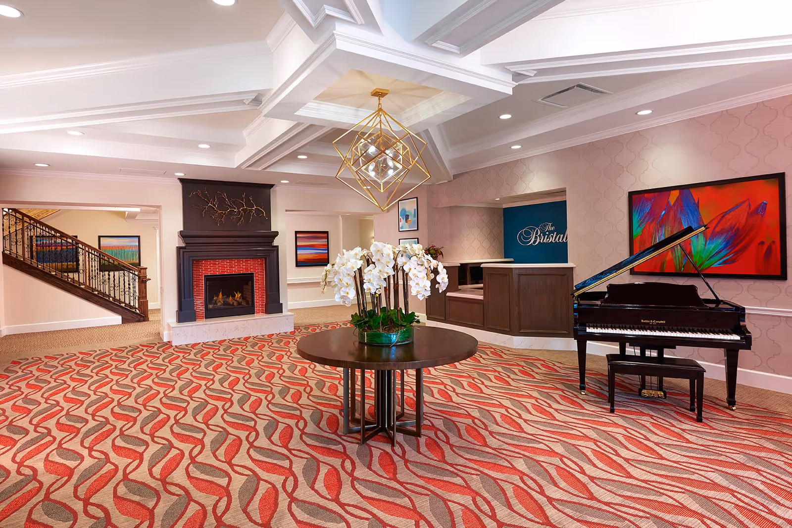 A spacious and elegant lobby area with a patterned red and beige carpet, a round wooden table with white orchids in the center, a black grand piano with a matching bench on the right, a fireplace with a black mantle and red brick interior on the left, colorful abstract paintings on the walls, a staircase with decorative railing in the background, and a reception desk with a sign that reads 'The Bristal'.
