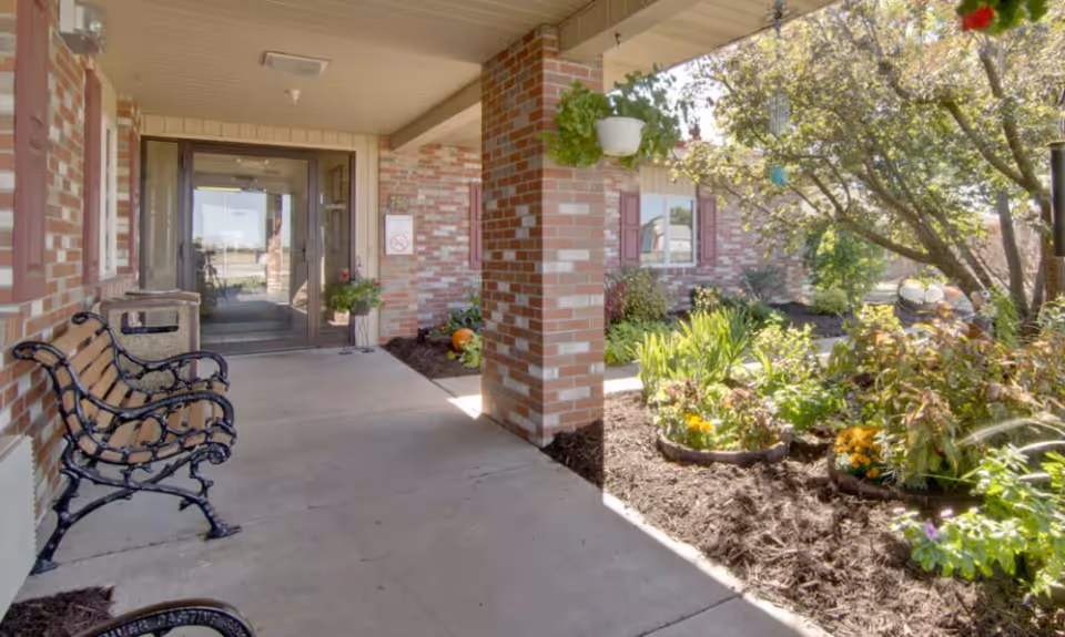 Covered brick entrance with benches, hanging planters, and a landscaped garden leading to glass double doors.