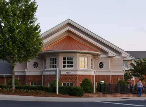 Exterior view of a senior living facility building with beige siding and red brick lower walls, featuring a large bay window with white trim and a copper-colored roof section. There is a tree on the left side and a person walking on the sidewalk on the right side.