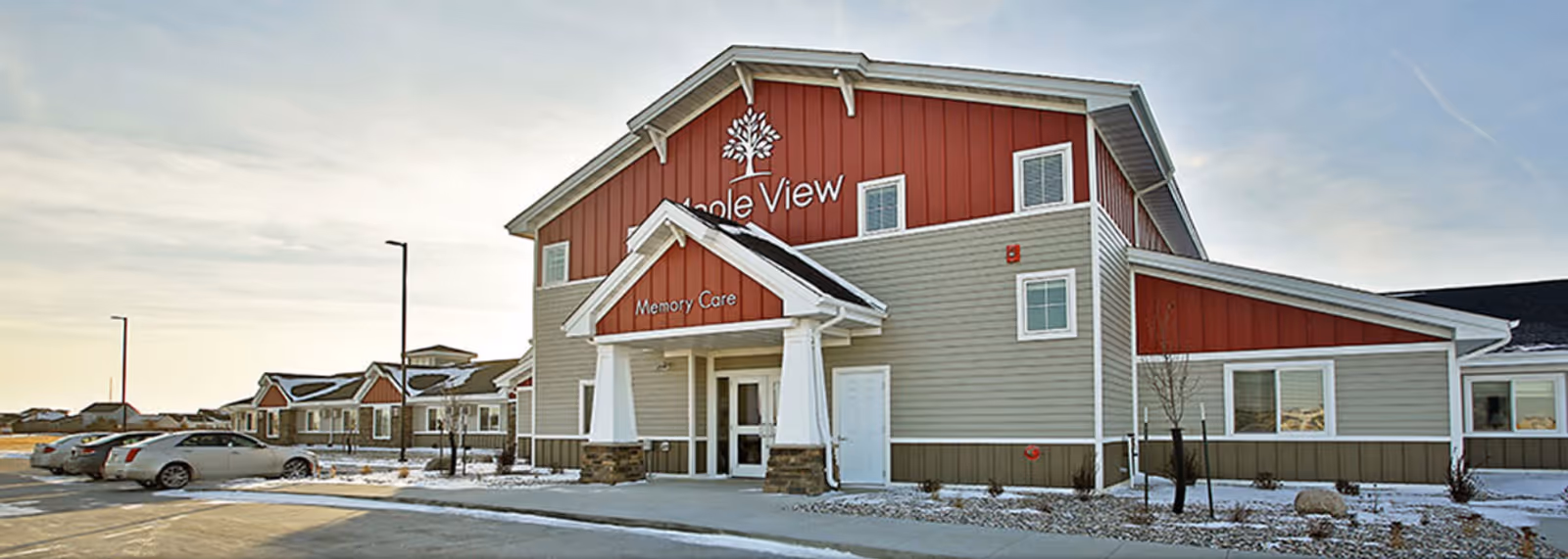Exterior view of Fargo Maple View senior living facility with a red and gray building labeled Memory Care. Several cars are parked in the parking lot in front of the building, and there is some snow on the ground.