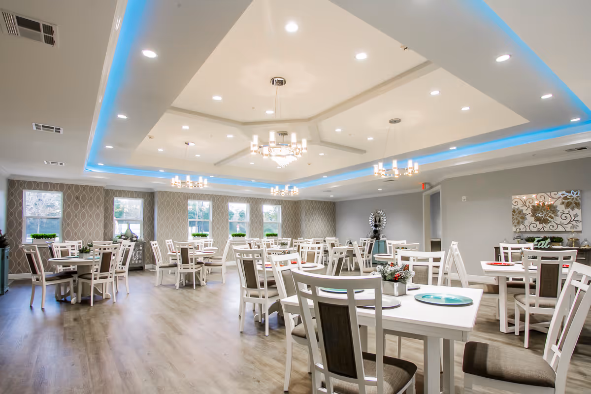 A spacious and well-lit dining room with multiple white tables and chairs arranged neatly. The room features large windows with patterned wallpaper on one wall, modern chandeliers hanging from a tray ceiling with blue accent lighting, and decorative plants on the tables. The floor is a light wood finish, and there is a decorative mirror and artwork on the far wall.
