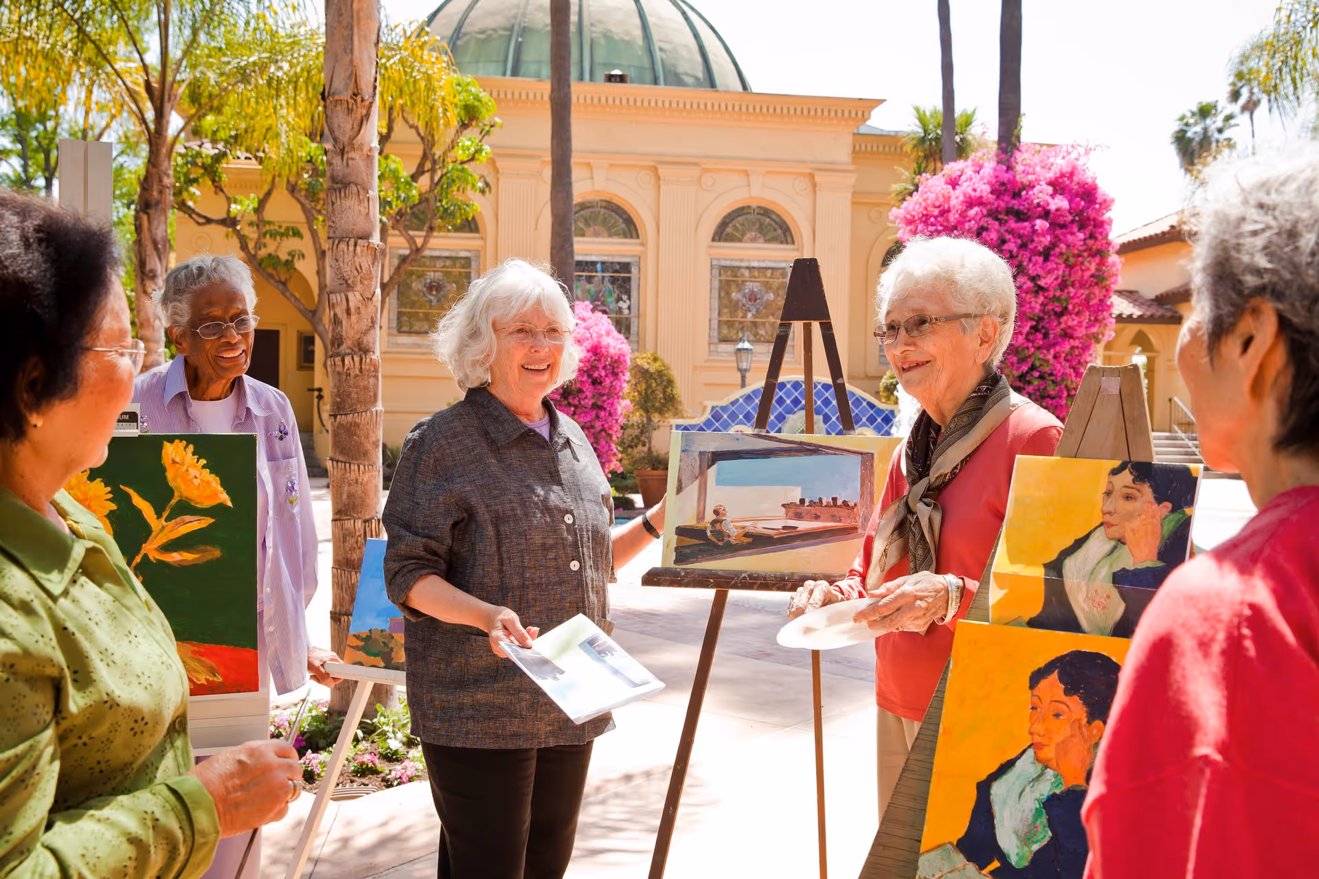 A group of elderly women standing outdoors in a sunny courtyard, smiling and engaging in an art activity with paintings displayed on easels around them. The background shows a building with arched windows, palm trees, and vibrant pink flowers.