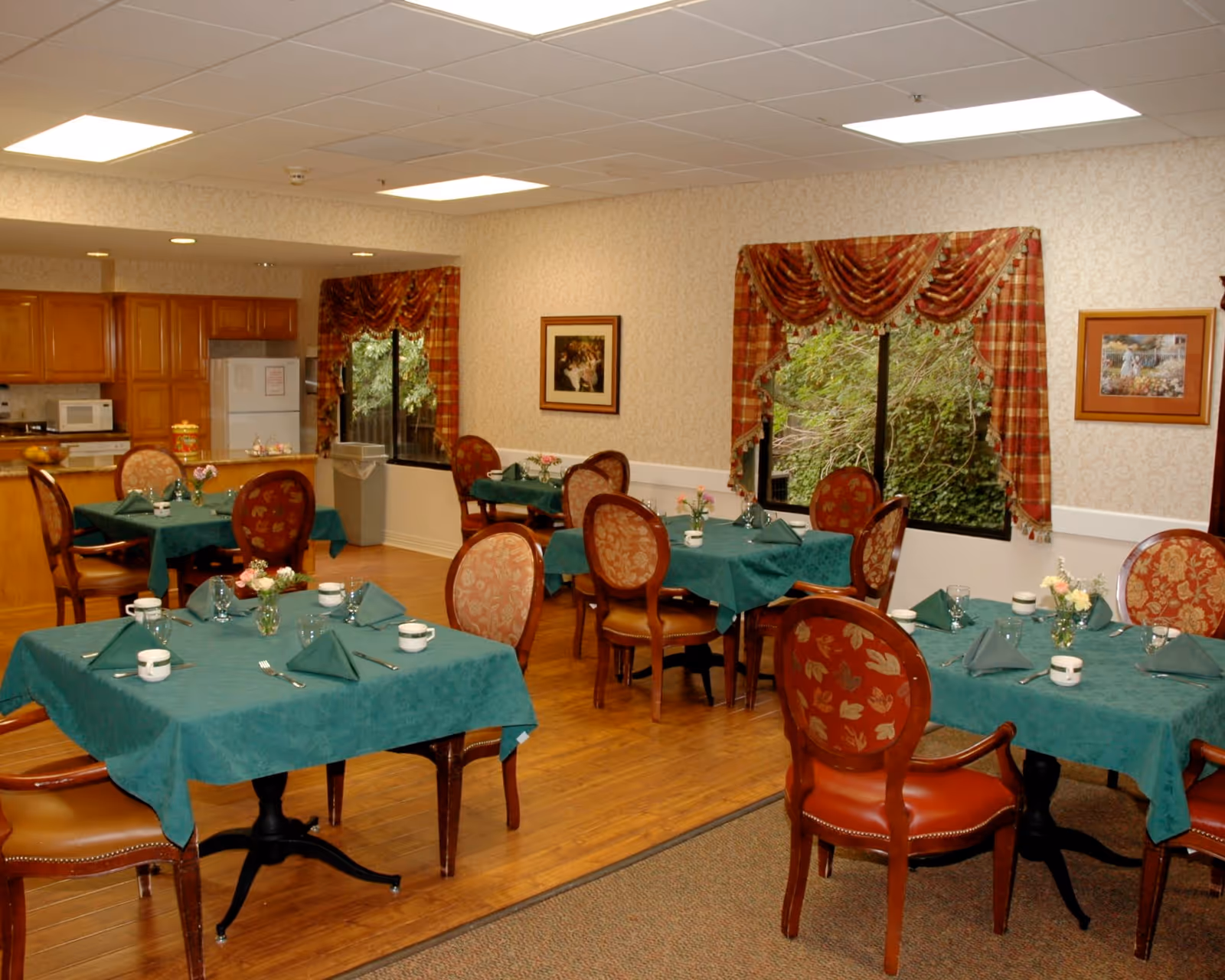 A dining room with several tables covered in green tablecloths, each set with folded napkins, cups, glasses, and silverware. The room has wooden flooring, patterned wallpaper, and windows with red and gold draped curtains. There are framed pictures on the walls and a kitchen area with wooden cabinets and appliances in the background.