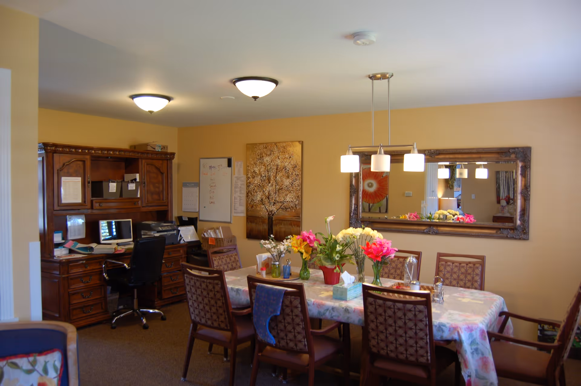 A dining room with a rectangular table covered with a floral tablecloth and surrounded by eight chairs. The table is decorated with several vases of colorful flowers and a tissue box. On the wall behind the table is a large ornate mirror reflecting part of the room and a painting of a flower. To the left, there is a wooden desk with a computer, printer, and office supplies. The walls are painted a warm beige color and the room is lit by ceiling lights and a hanging light fixture above the table.